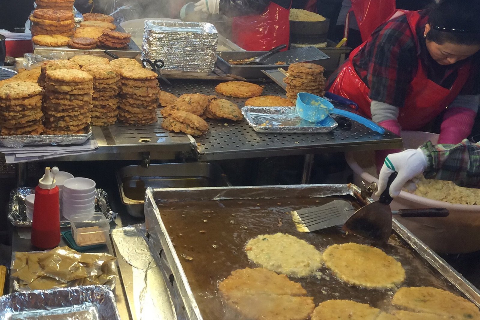 Mung bean pancakes frying on a large griddle beside stacks of finished pancakes