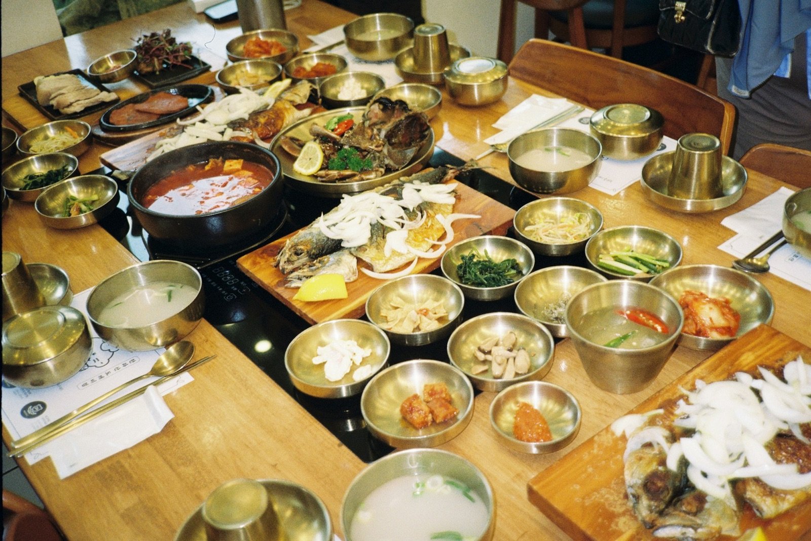 A wooden table covered with small brass bowls of Korean side dishes and stew
